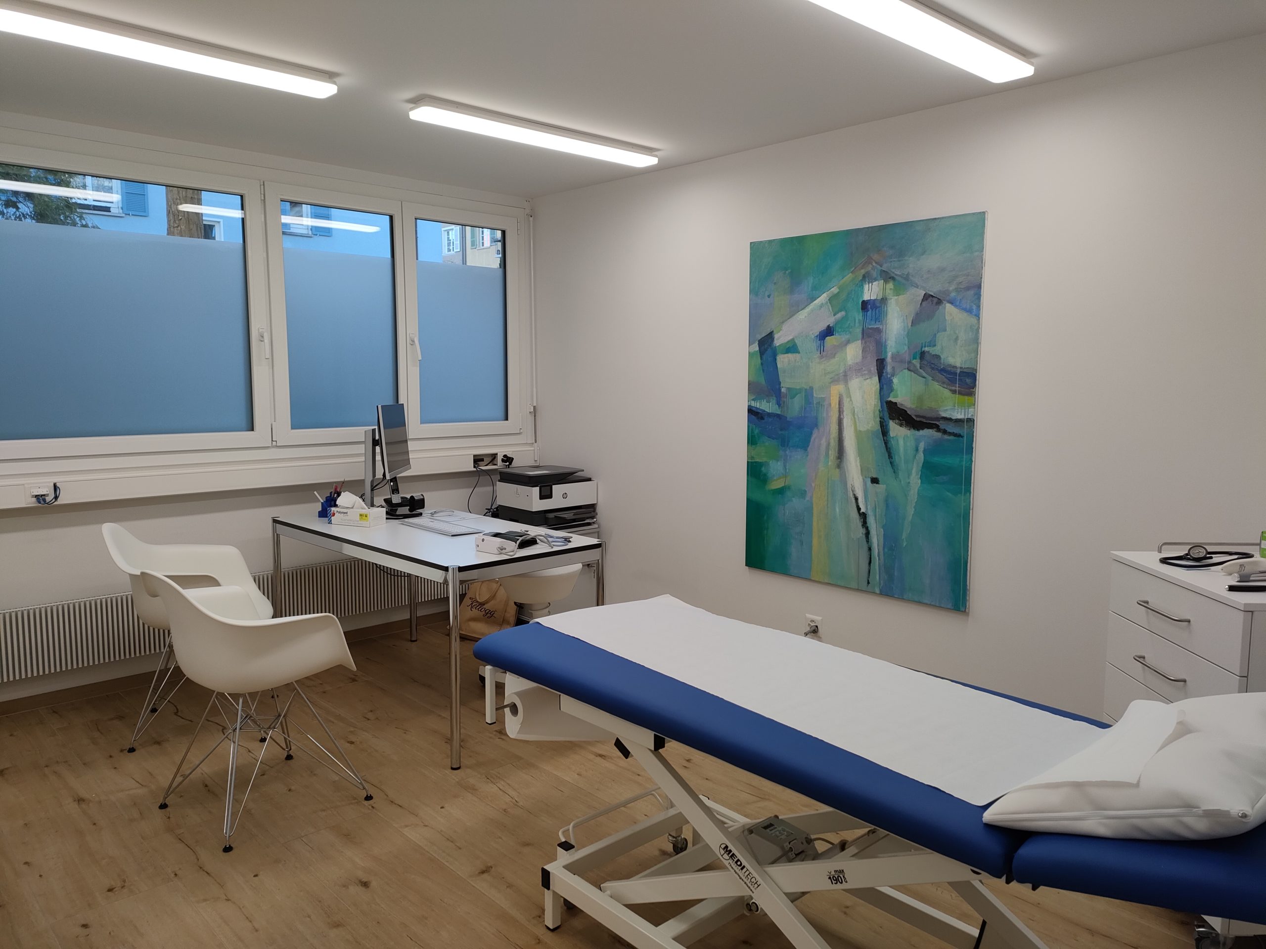 Ein Patientenzimmer mit einem blau-weissen Bett, Schreibtisch, Stühle und ein Gemälde an der Wand.